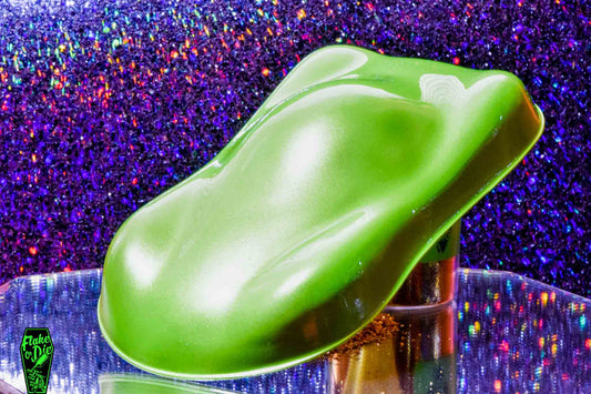 Macro photography of a car like speed shape painted with lime green pearl pigment. The lighting contrasts dark and light lime green on the curves of the shape. Mirrored platform and complementary background of blue/rainebow bokeh.