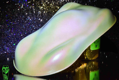 Macro photography of a car like speed shape painted with white colorshift pearl pigment. Lighting contrasts of white, purple, pink and green on the curves of the shape. Mirrored platform and complementary background of silver bokeh.
