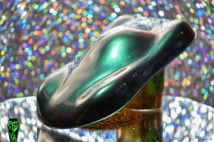 Macro photography of a car like speed shape painted with green/gold pearl pigment. Lighting contrasts of dark and light greens with gold glimmer on the curves of the shape. Mirrored platform and complementary background of rainbow bokeh.