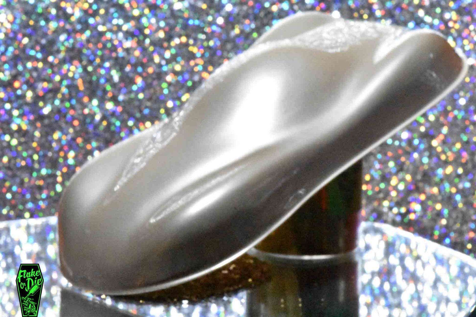 Macro photography of a car like speed shape painted with silver pearl pigment. The lighting contrasts dark and light silver on the curves of the shape. Mirrored platform and complementary background of rainbow bokeh.