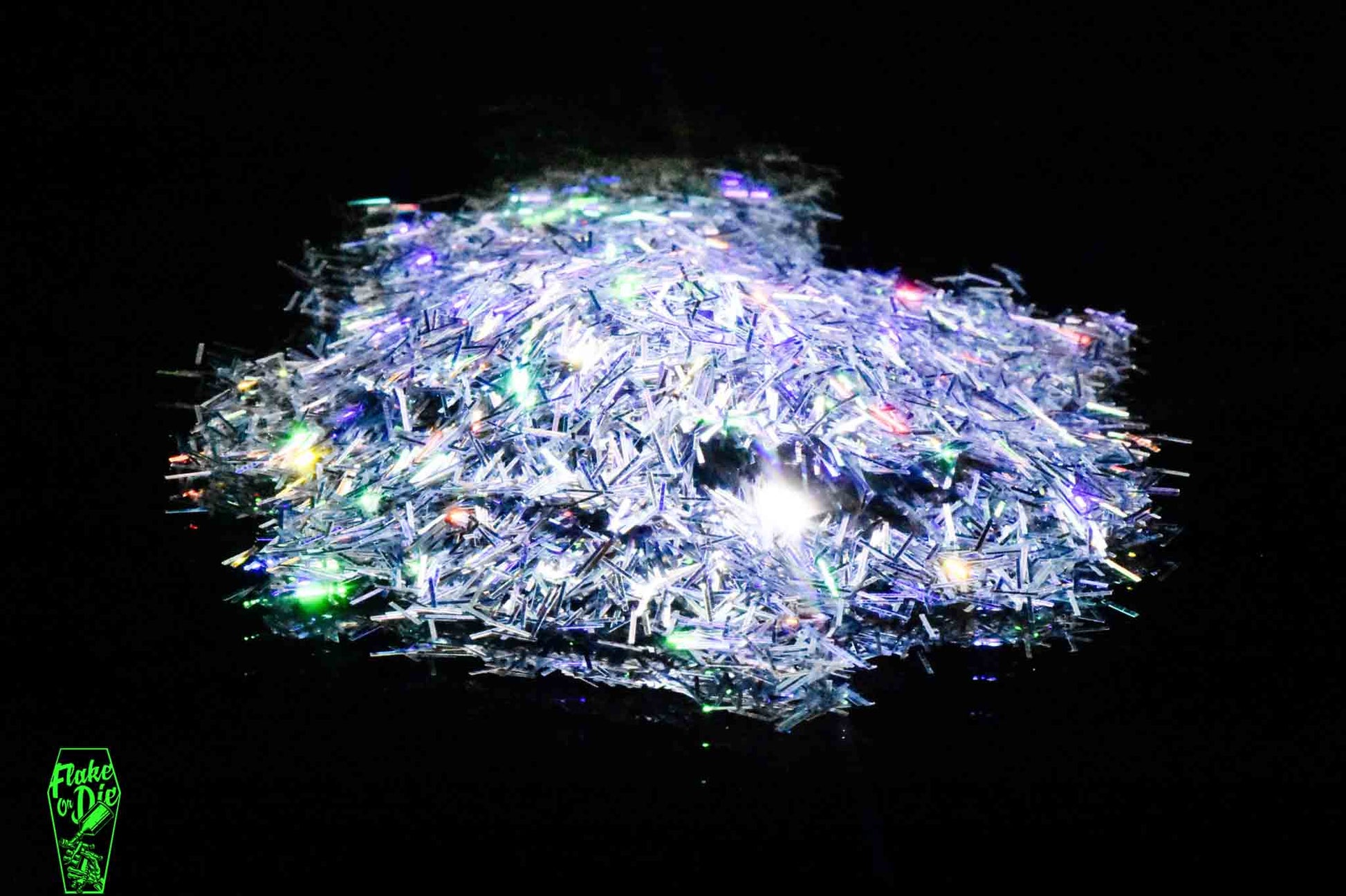 Macro photography of a small pile of silver holographic splinter cut metal flake particles. Distinct glitter flares of yellow, green, red and orange on the metal flake and a stark black background.